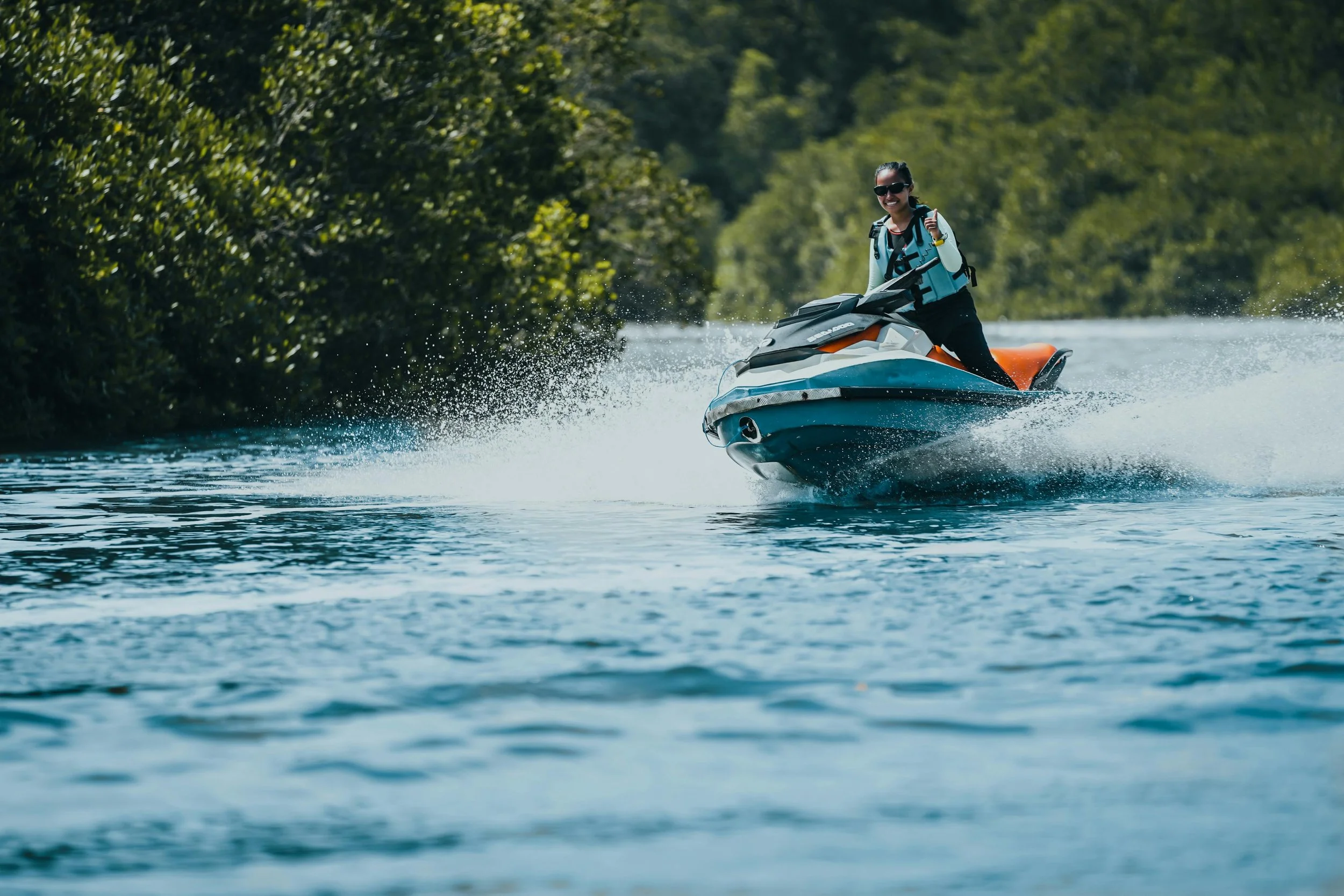 A woman riding a jet ski on a waterway surrounded by trees, smiling and wearing sunglasses and a life vest.