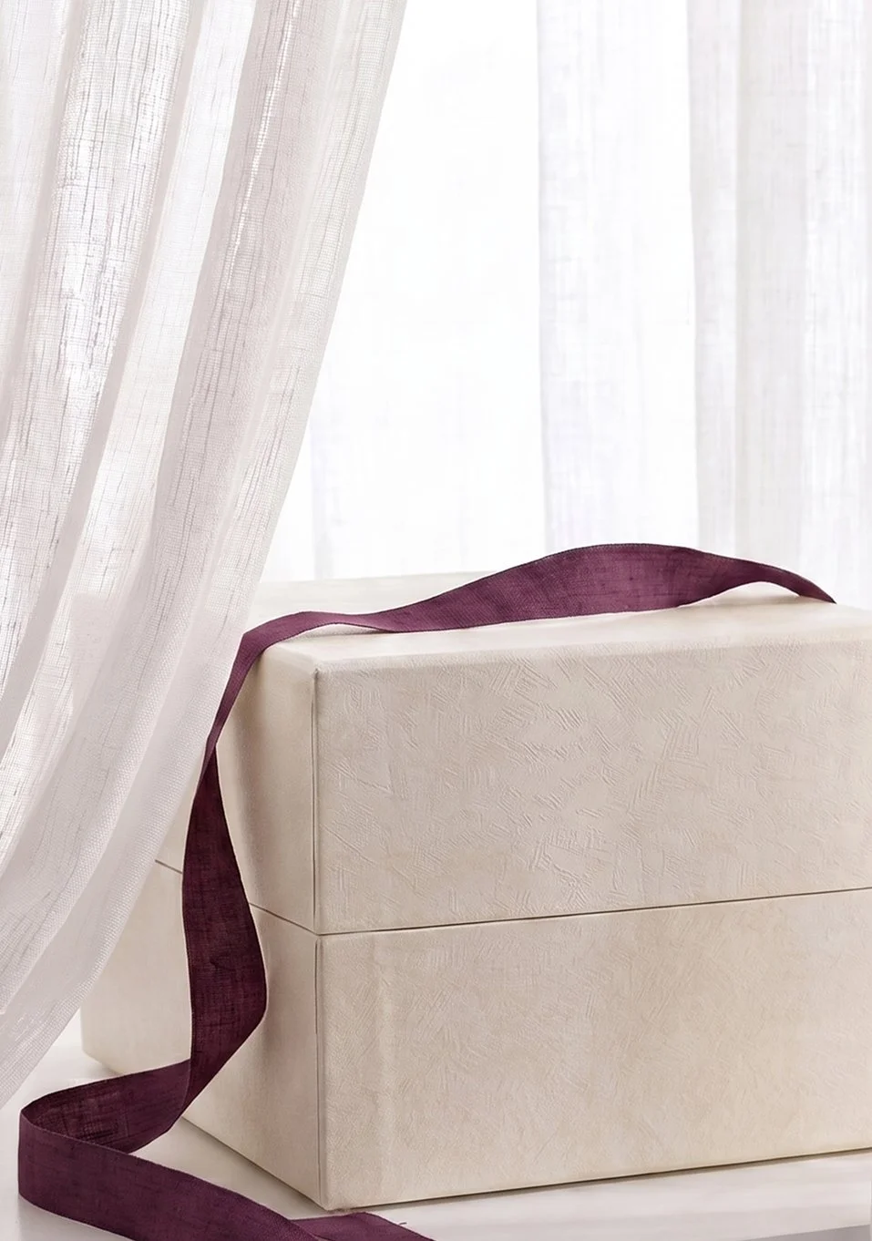 A gift box with a purple ribbon draped over it, partially covered by sheer white curtains.