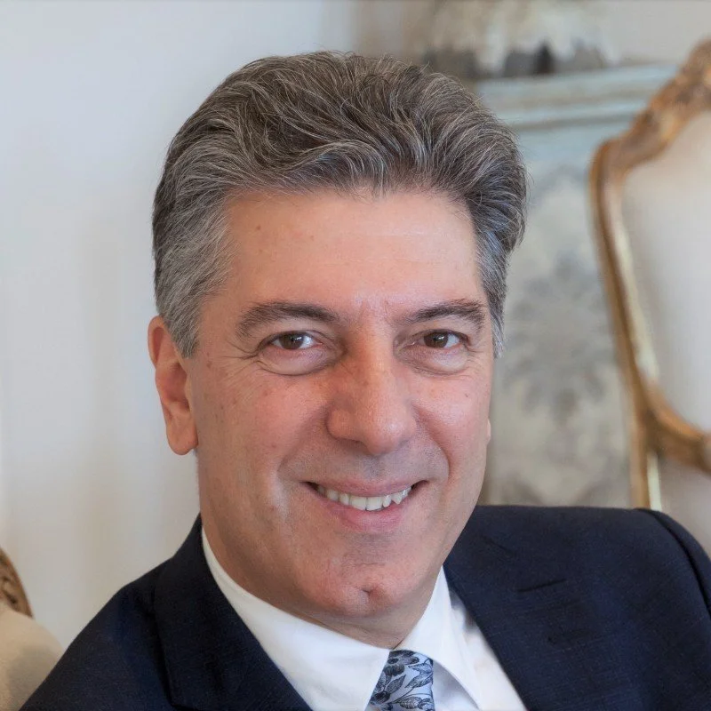 A smiling middle-aged man with gray hair, wearing a suit with a white shirt and a patterned tie, sitting in a room with classic decor.