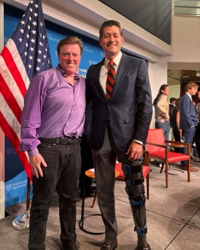 Two men standing side by side at a political event, one wearing a lavender dress shirt and the other in a suit with a patterned tie, with an American flag and a blue backdrop behind them. The man on the right has a knee brace.