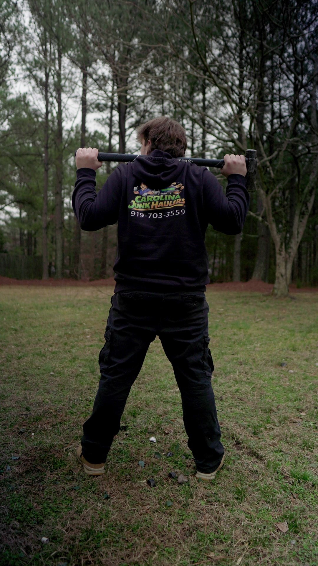 Person standing on grass, holding a sledgehammer behind their shoulders, facing away. Wears black hoodie with colorful logo and phone number, cargo black pants and tan shoes. Surrounded by trees in a wooded area.