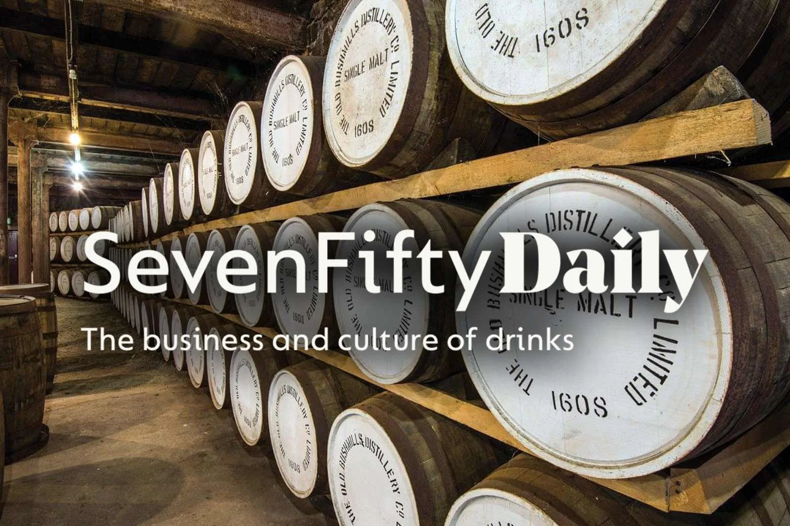 Rows of wooden barrels stacked on shelves in a distillery cellar, with branding and text on the barrelheads.