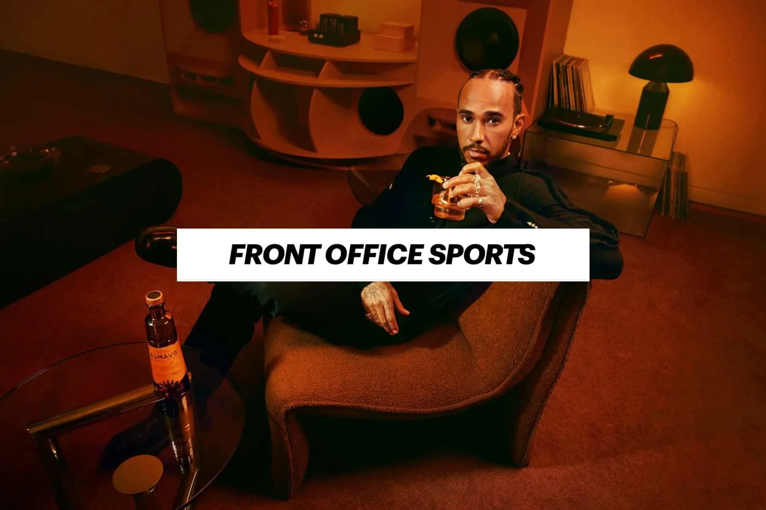 A man with braided hair and tattoos reclines on a vintage armchair in a dimly lit room, holding a glass in one hand, with a bottle of alcohol on a small table nearby. The room has warm lighting and modern decor, including a bookshelf and a lamp. Text overlay reads "FRONT OFFICE SPORTS."
