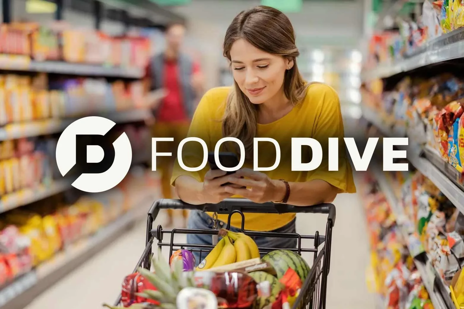 Woman shopping for groceries in a supermarket, looking at her phone with a shopping cart filled with bananas, watermelon, and other fruits.