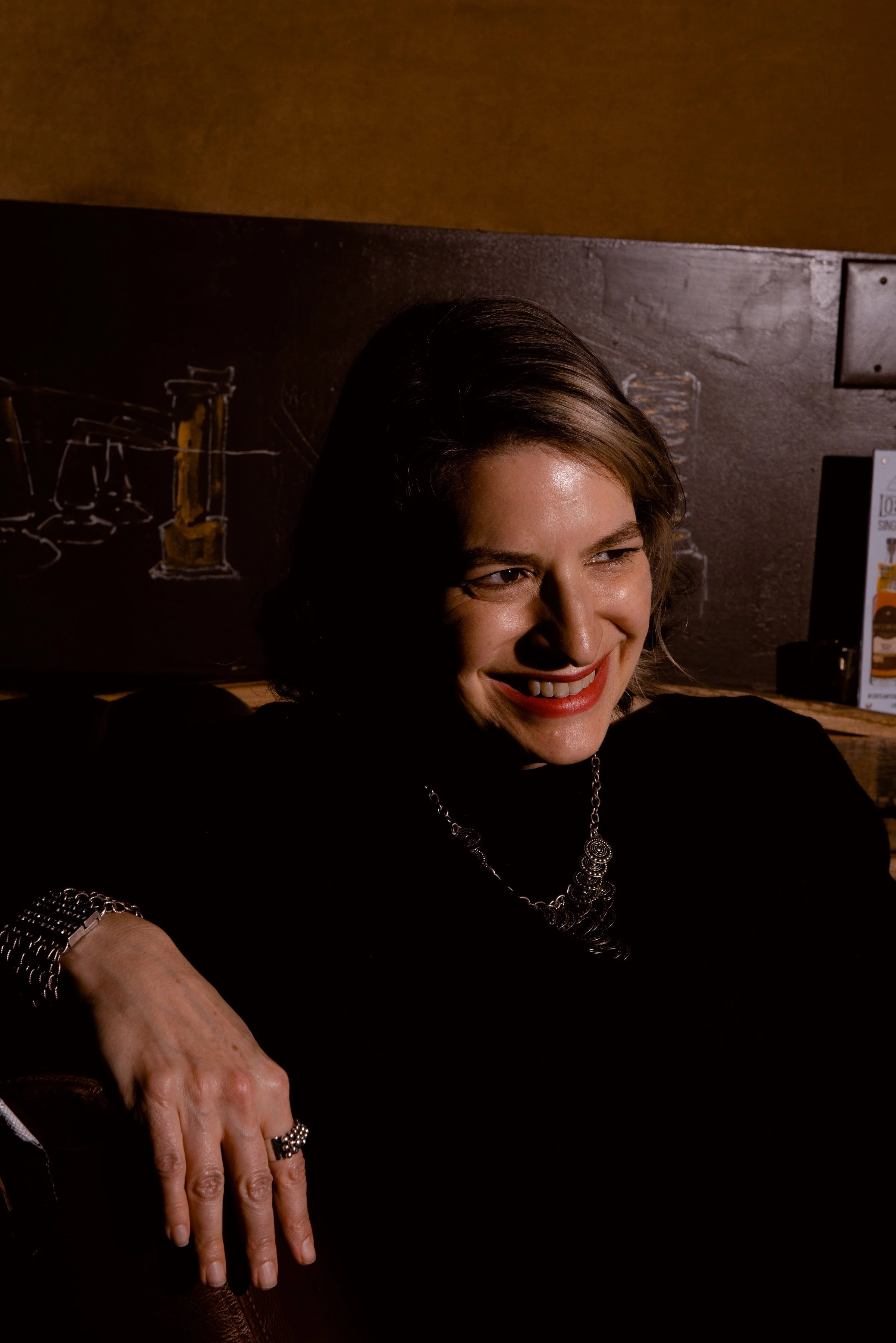 A woman with short dark hair smiling and wearing a black top, jewelry, and lipstick, sitting in a dimly lit setting with a chalkboard in the background.