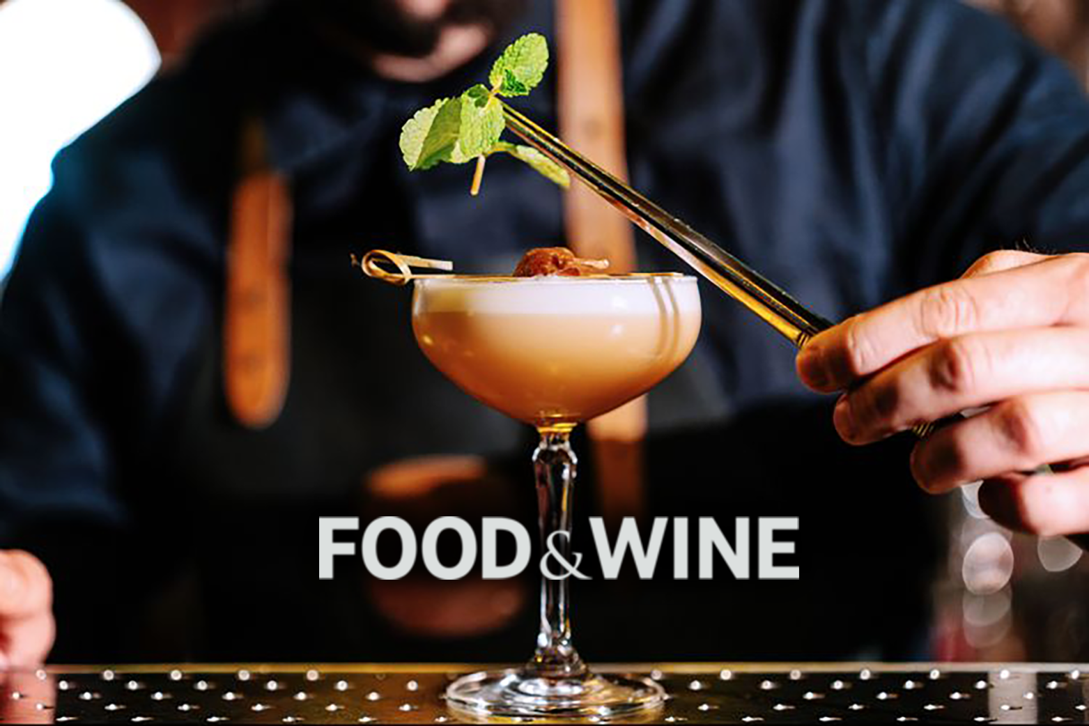 A bartender garnishing a cocktail with a sprig of mint using tongs. The cocktail is in a stemmed glass on a bar counter. The person is wearing a black shirt or apron. The words "FOOD & WINE" are displayed at the bottom of the image.