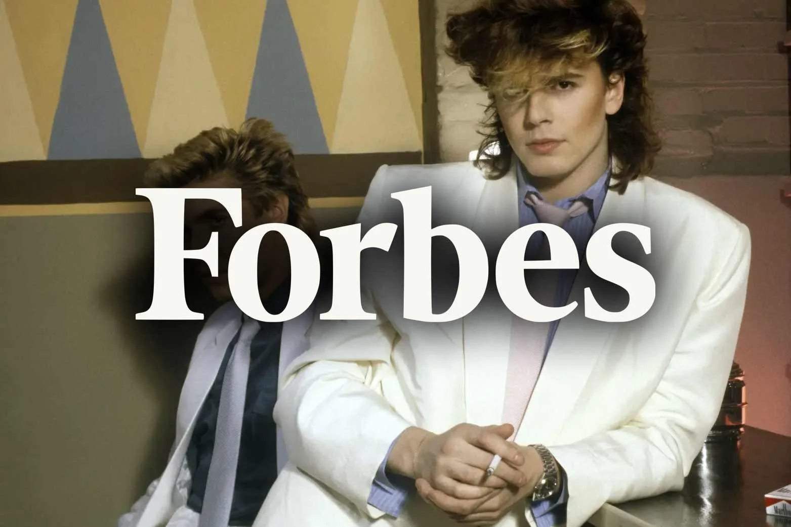 Two men sitting on a bench in a restaurant or bar. One man, on the right, is wearing a white suit and has a serious expression. The other man is partially obscured and is wearing a jacket and tie. The FORBES logo is overlayed on the image.