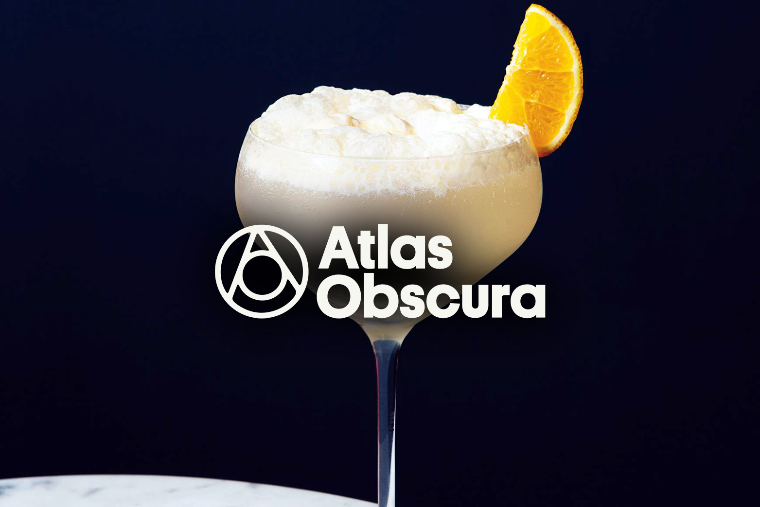 A cocktail glass filled with a creamy beverage topped with whipped cream and garnished with a lemon wedge on a dark background, with the text 'Atlas Obscura' overlayed.