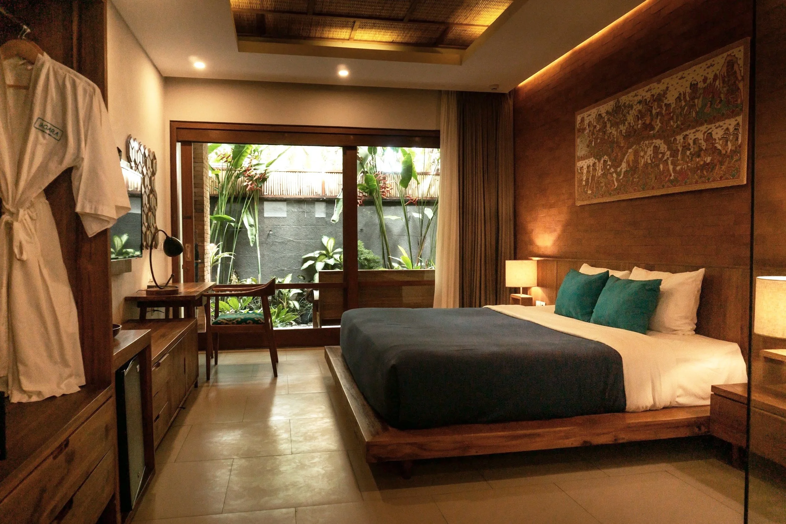 Hotel room with a large bed, wooden furniture, a balcony with tropical plants, and warm lighting.