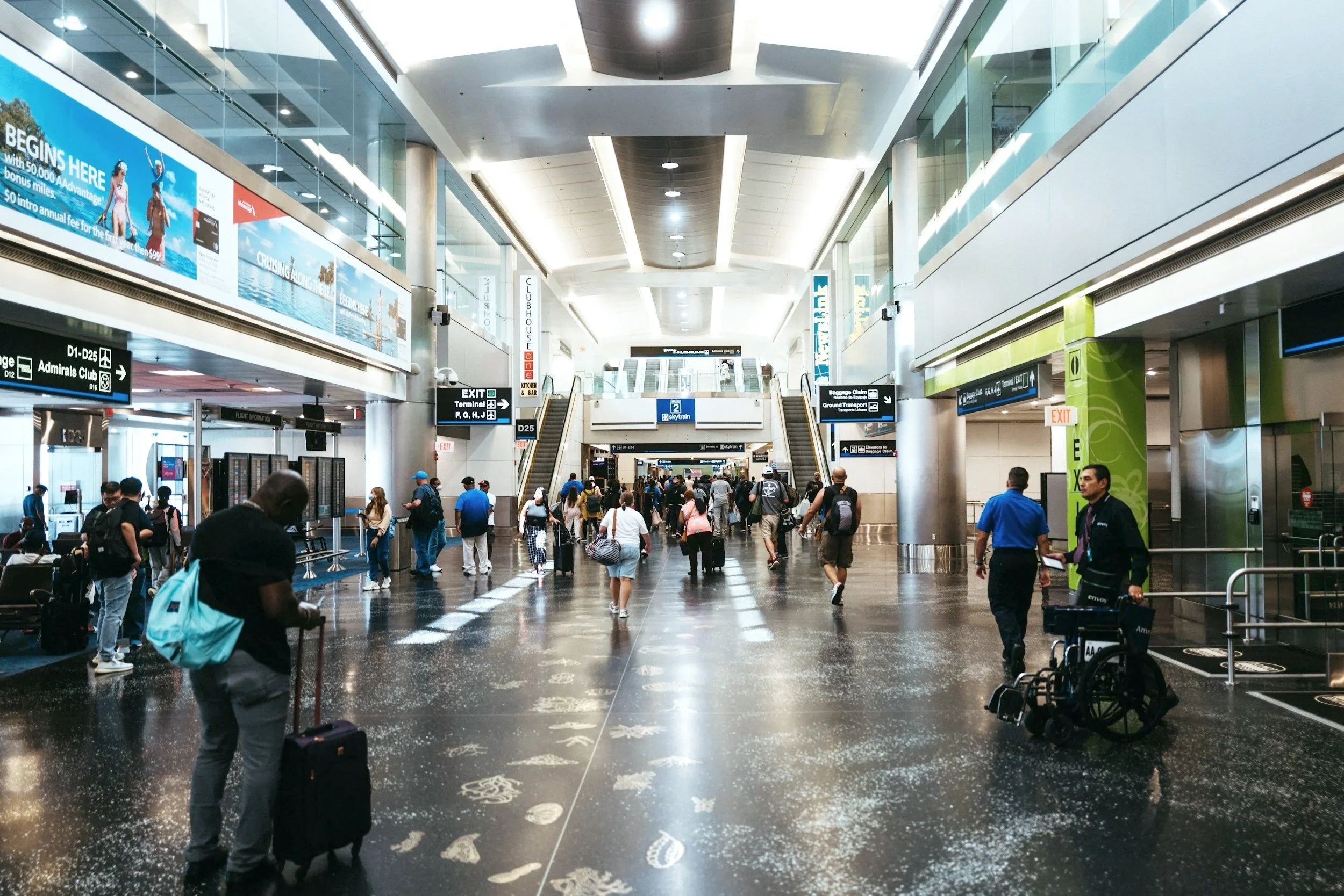 Interior of an airport terminal with travelers walking and standing, signage indicating gates and exits, escalators, and polished black floor with footprints.