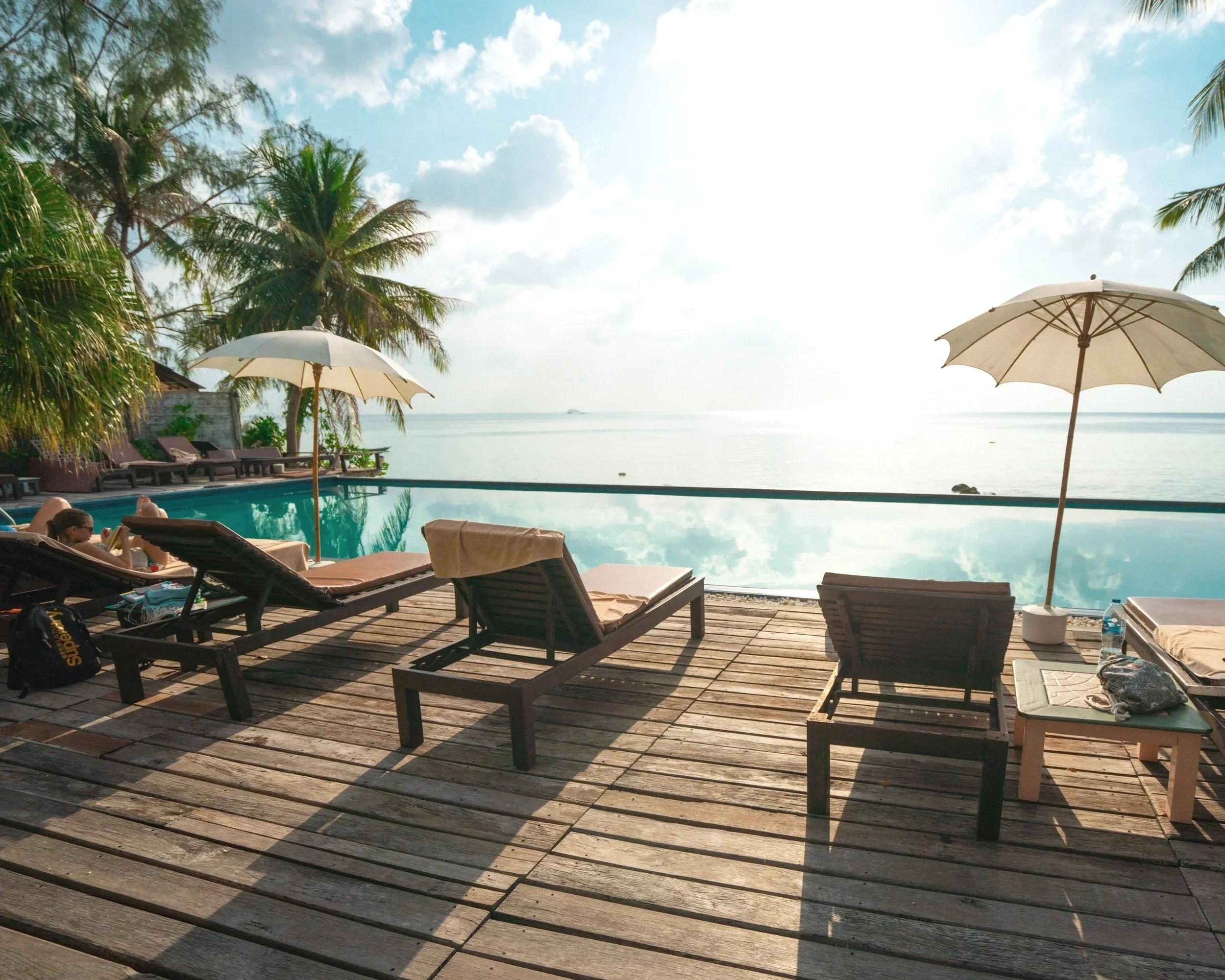 Poolside views with lounge chairs, umbrellas, palm trees, water, and a clear sky.