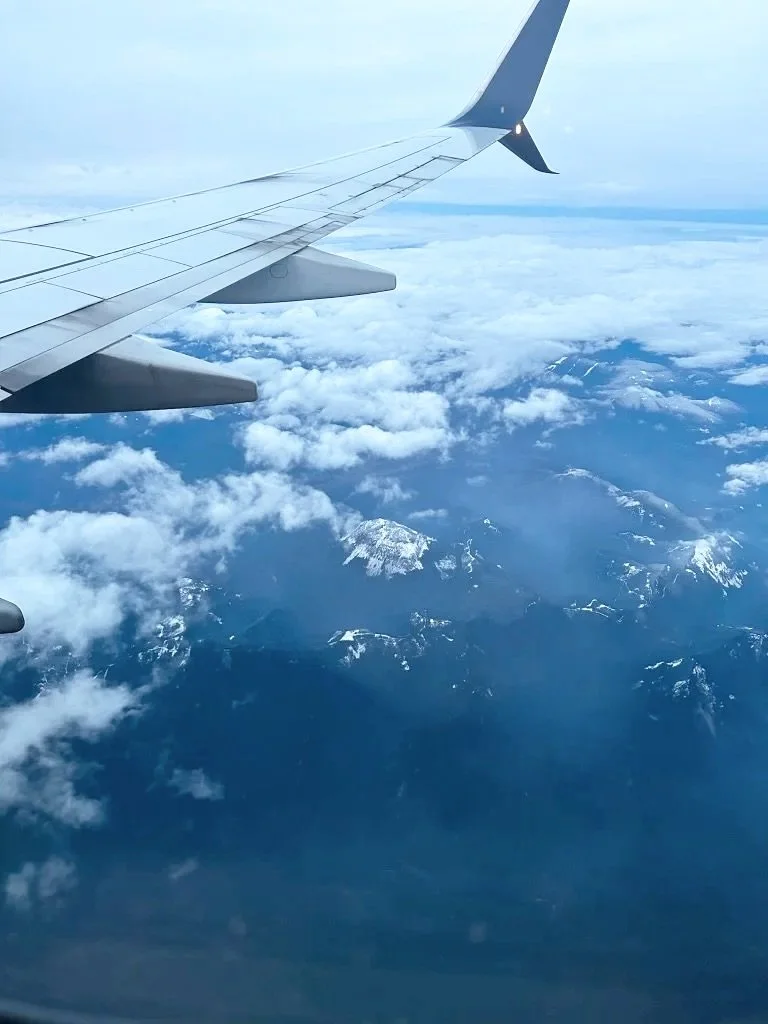 View from an airplane window showing part of the wing and clouds below, with snow-capped mountains in the distance.