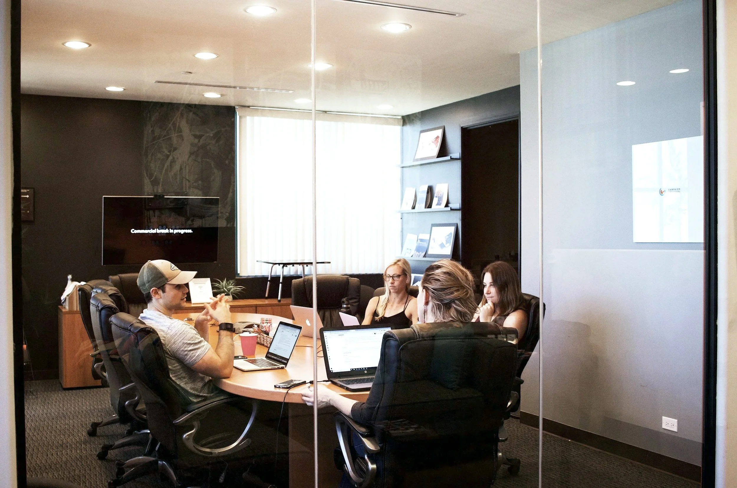 Four people in a conference room having a meeting, with laptops and drinks on the table, viewed through glass walls.