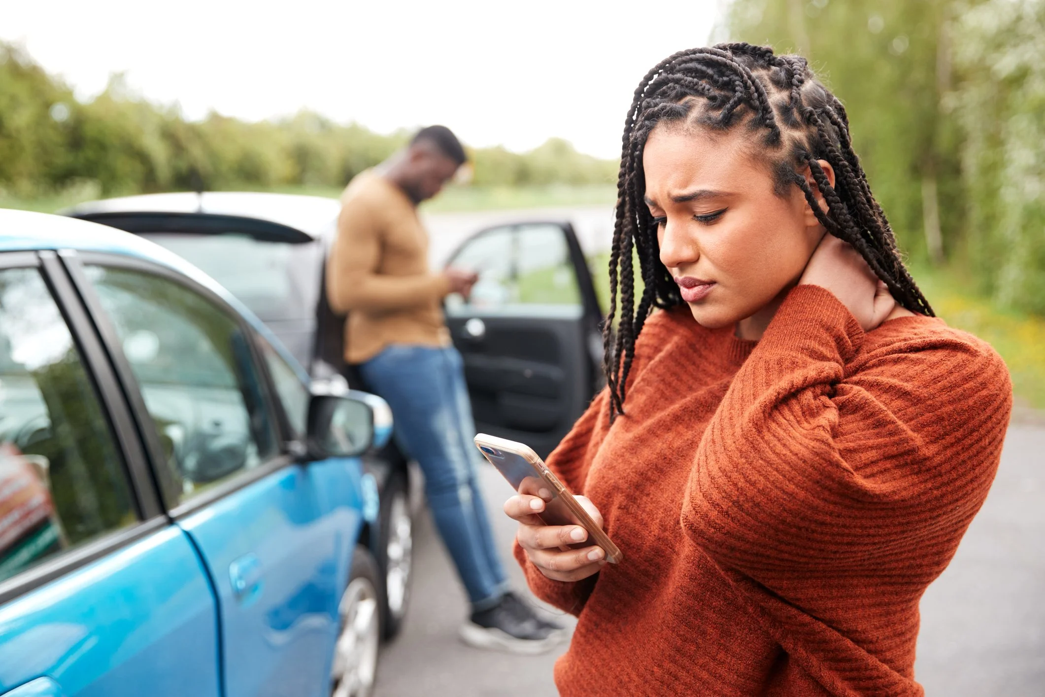 A distressed woman with braided hair and wearing an orange sweater holds her neck and looks at her phone near two parked cars on a roadside; a man in the background looks at his phone beside his car.