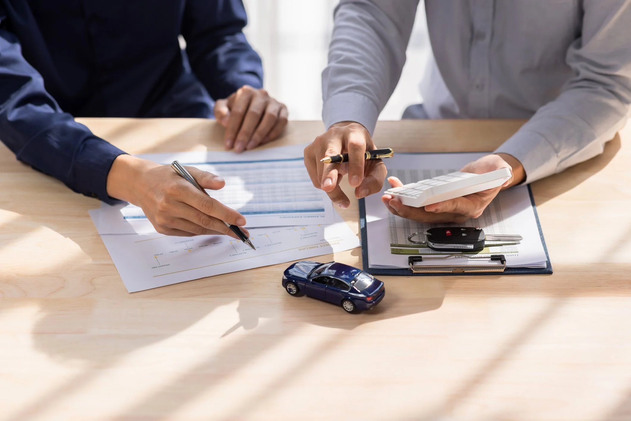 Two people reviewing documents on a desk, with a small blue model car, car keys, and a calculator on the table.