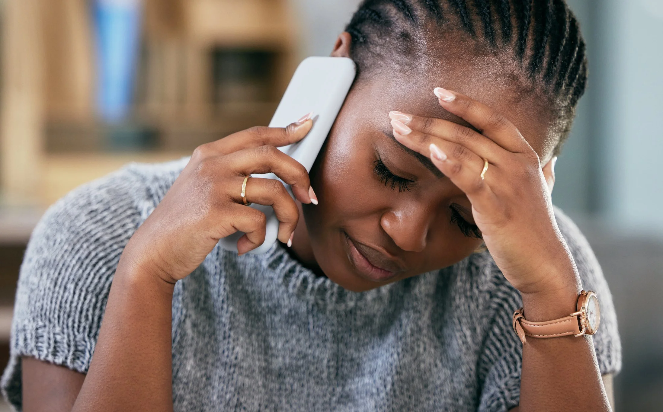 A woman with braided hair, wearing a grey patterned top, is holding her forehead with one hand and talking on a mobile phone with the other, appearing stressed or worried.