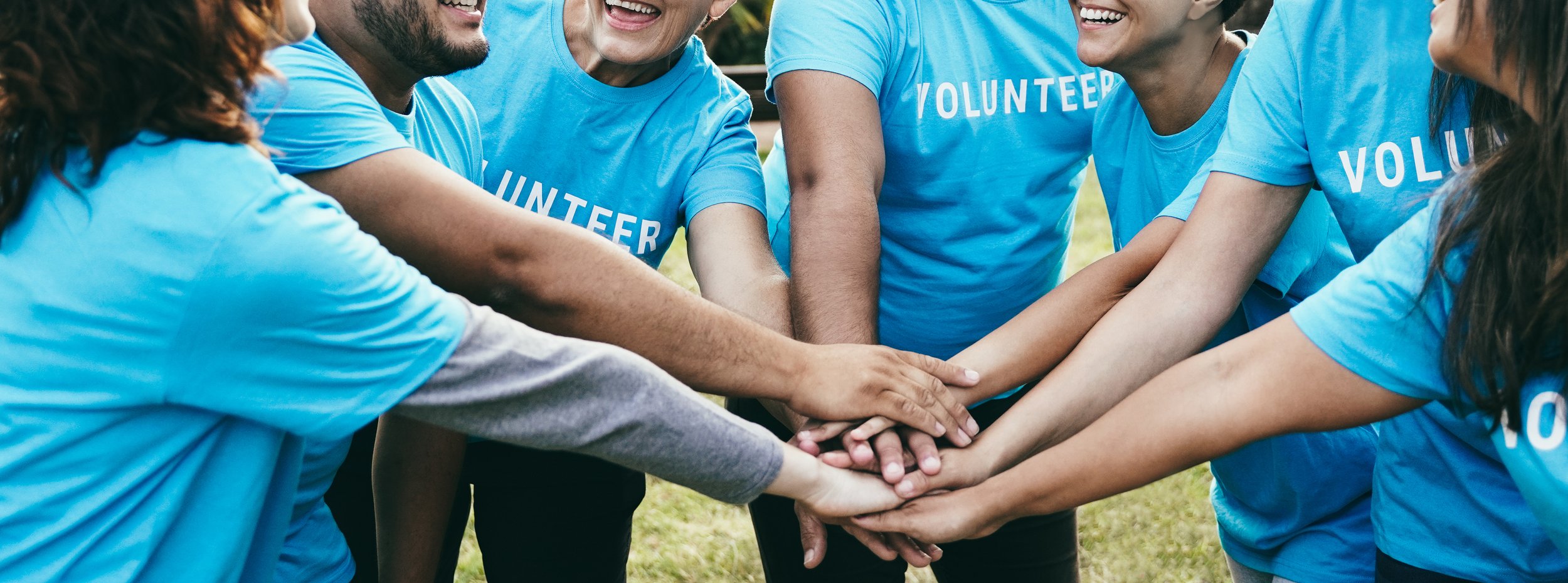 A group of volunteers wearing blue shirts with the word 'VOLUNTEER' printed on them, standing outdoors, putting their hands together in a show of unity.