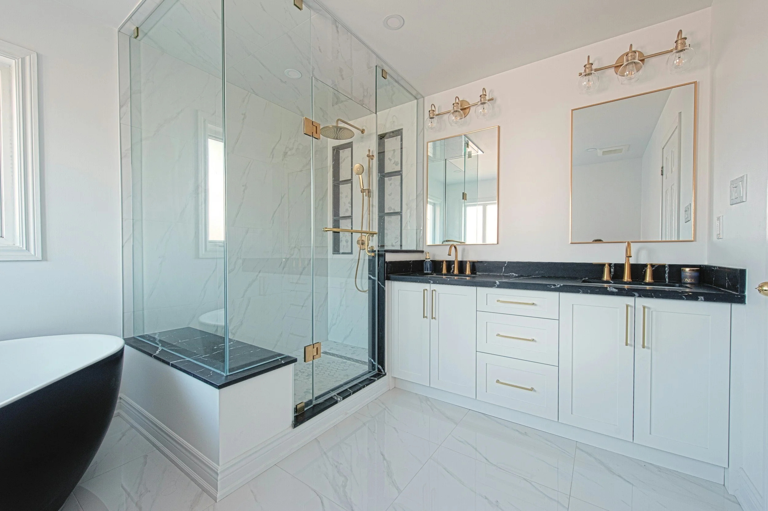 Modern bathroom with a glass-enclosed shower, white cabinetry, black marble countertop, gold fixtures, and double mirrors.