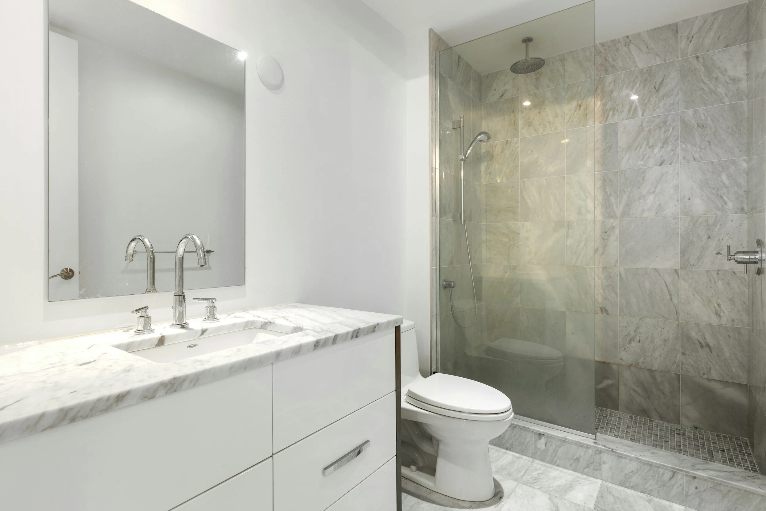 Modern bathroom with marble vanity, large mirror, toilet, and glass-enclosed shower with marble walls and ceiling-mounted showerhead.