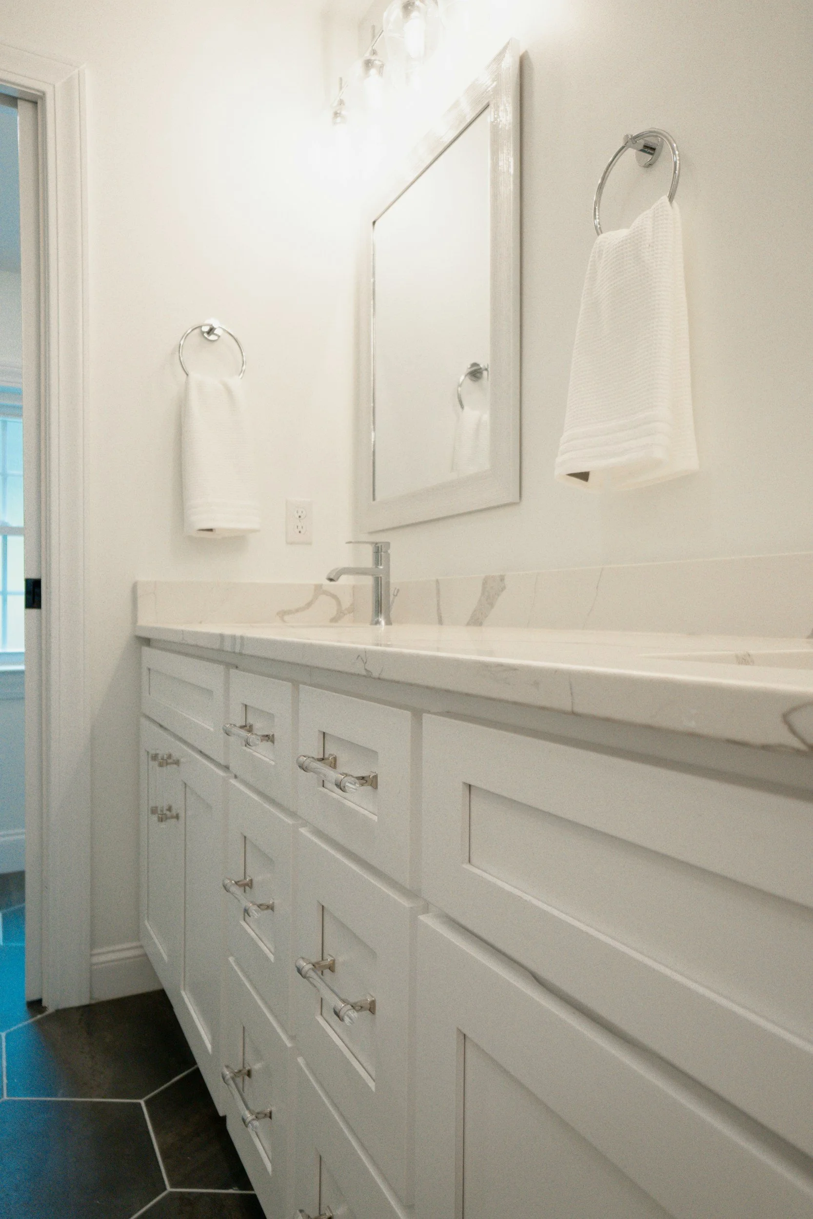 Bathroom vanity with white cabinets, marble countertop, and a square mirror. Two white hand towels hang on chrome rings on the wall. There is a faucet and a white wall outlet. Part of a door frame and partial view of a window are visible.