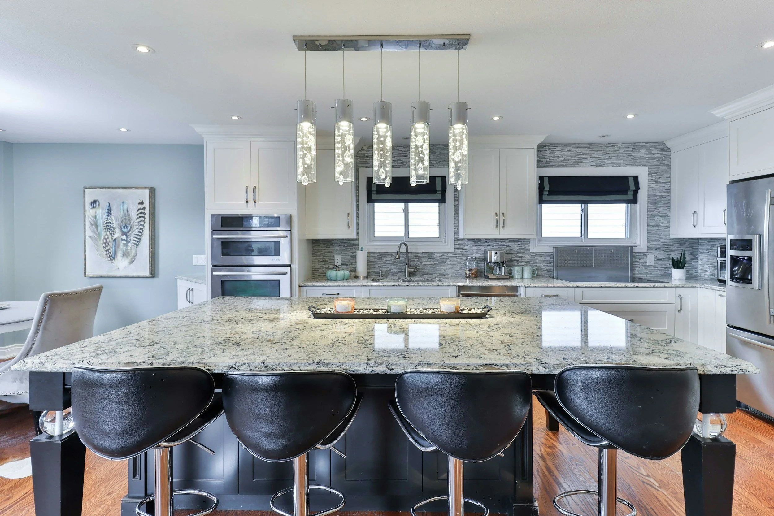 Modern kitchen with white cabinets, granite countertops, stainless steel appliances, a central island with black stools, pendant lighting, and dual windows with black blinds.