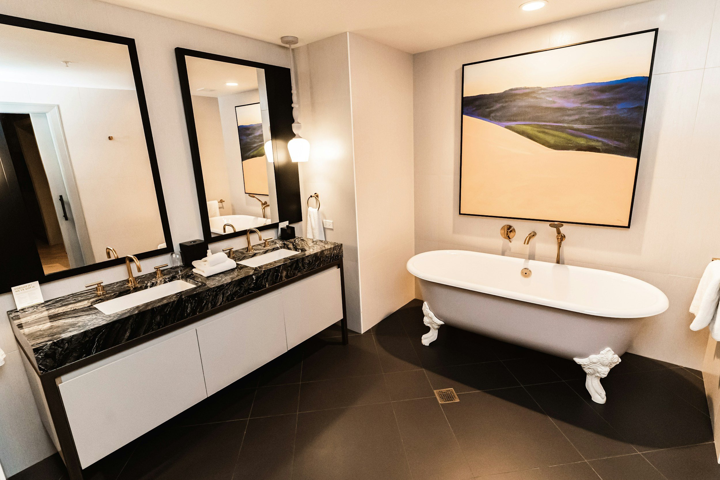 Modern bathroom with black marble double vanity, large mirrors, white bathtub with ornate clawfoot legs, and abstract landscape painting on the wall.