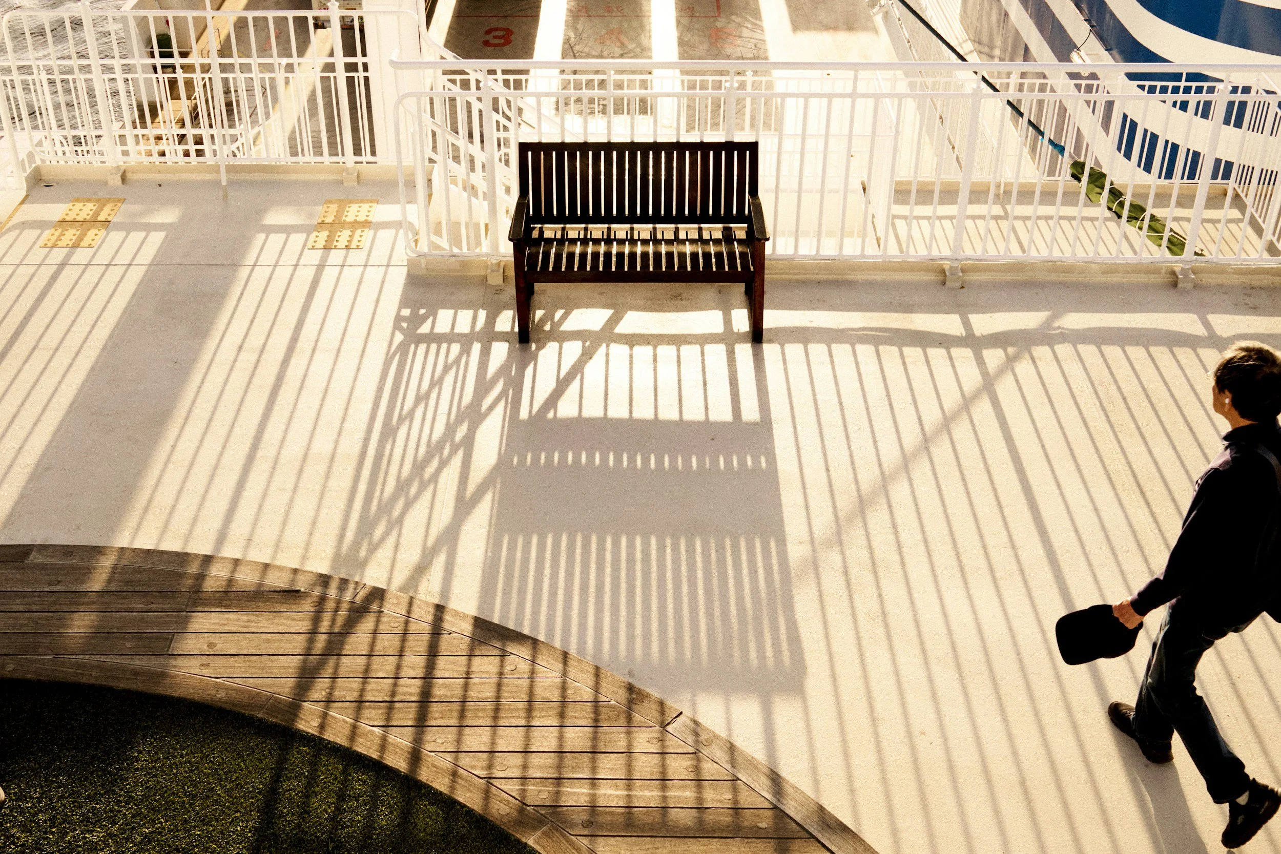 A man walking on a balcony holding a black hat, with shadows of railing lines cast on the ground and a brown bench nearby.