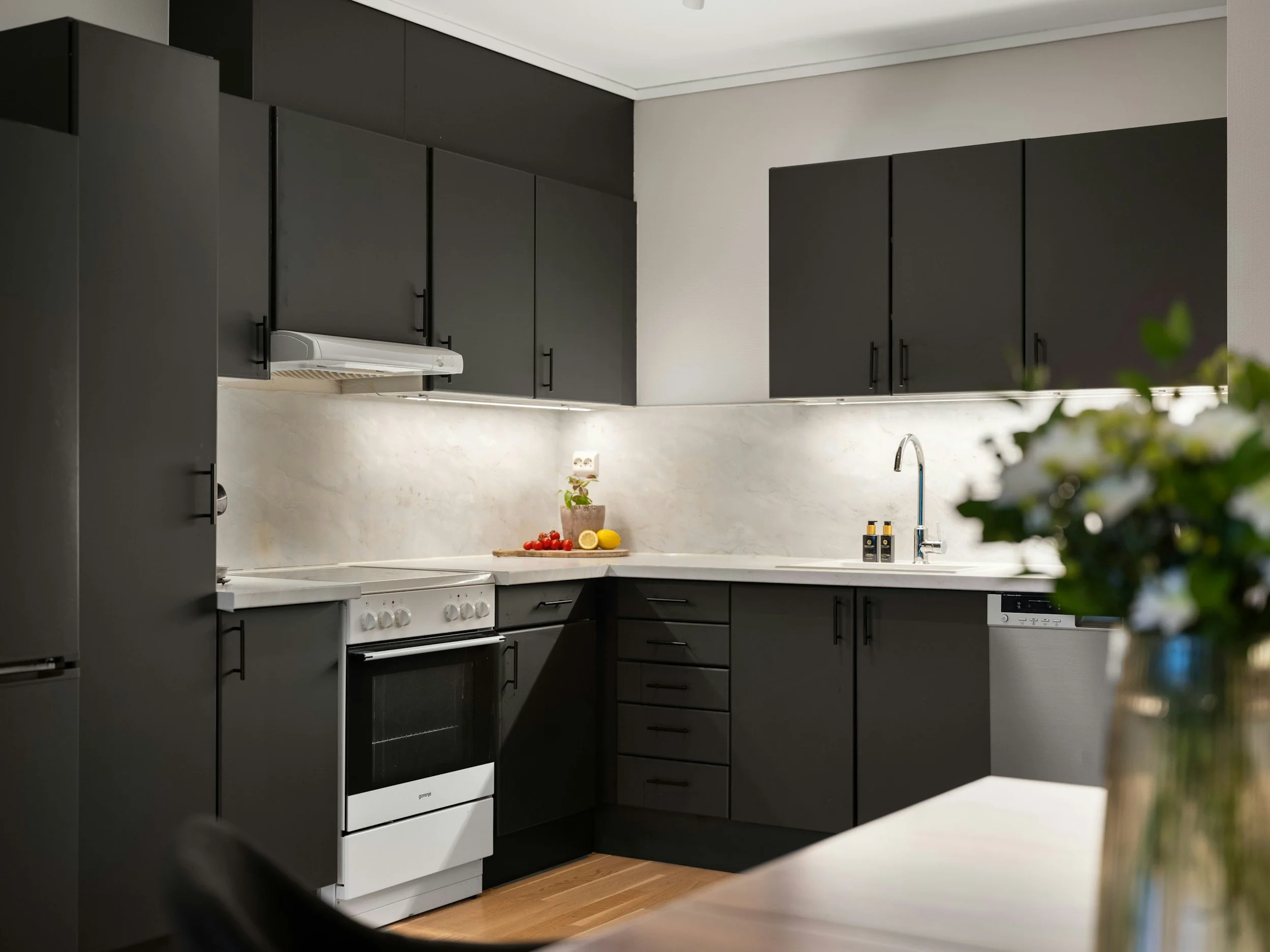 Modern kitchen with black cabinets, white countertops, a white stove, and a sink. There's a vase with flowers in the foreground, and a few bottles and some fruits on the counter.
