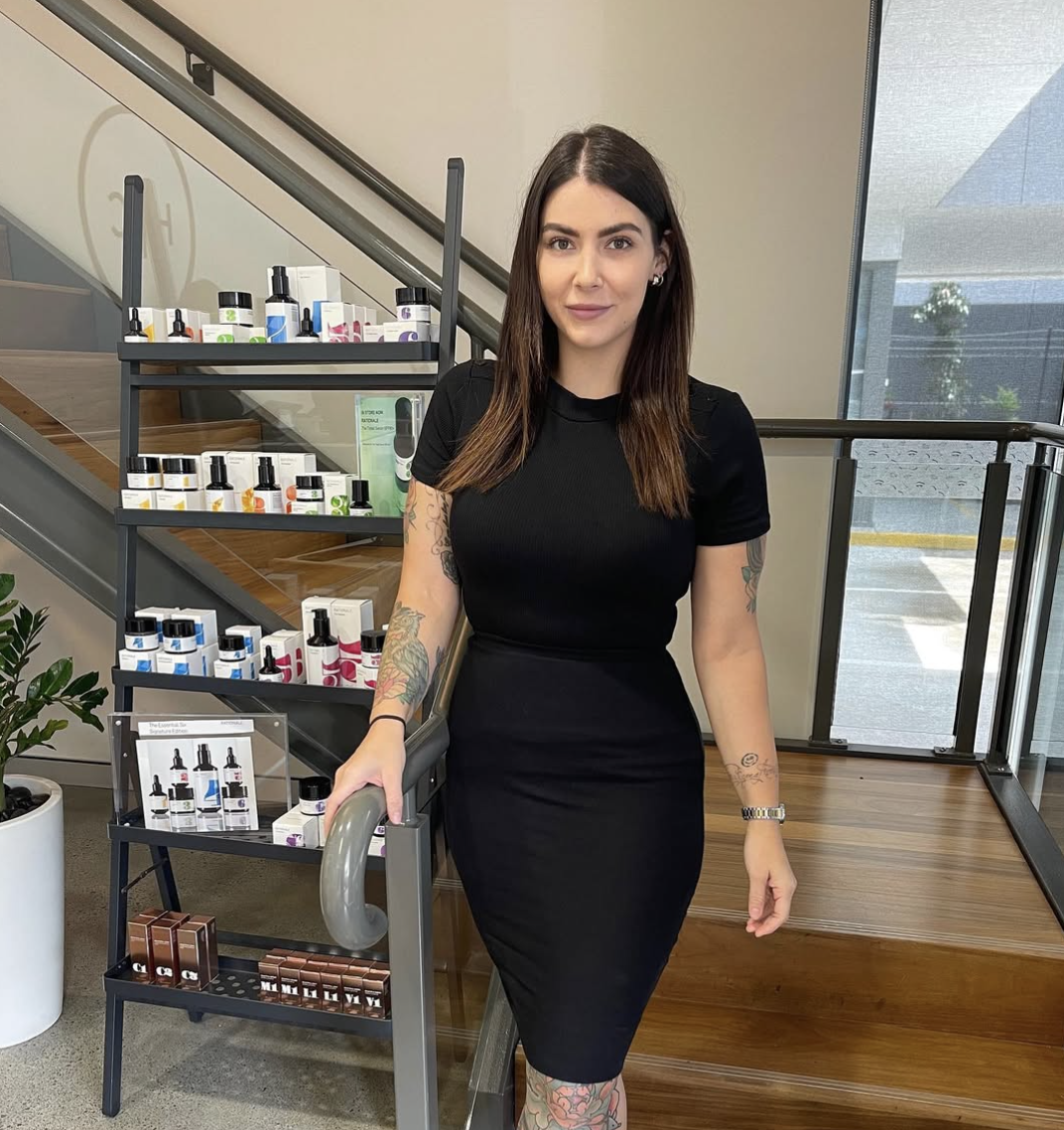 Martika Bennett, Founder of Sana Skin Clinic, wears a fitted black dress standing near stairs with skincare products displayed on shelves behind her.