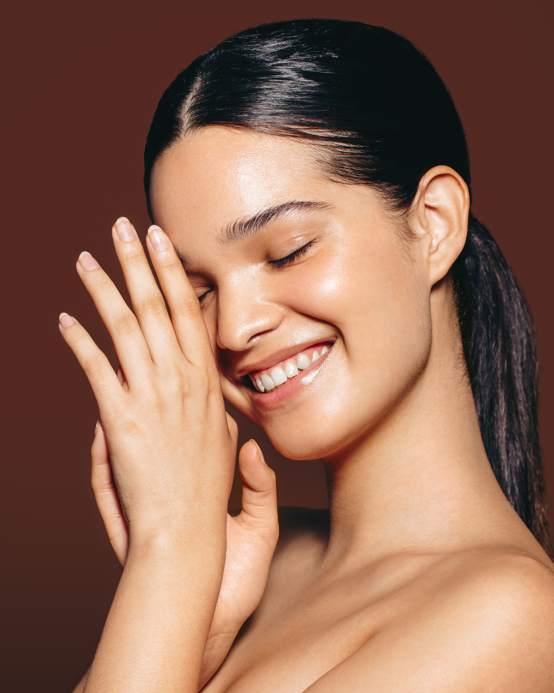 A woman with dark hair styled in a ponytail, smiling with eyes closed and one hand touching her face, against a brown background.