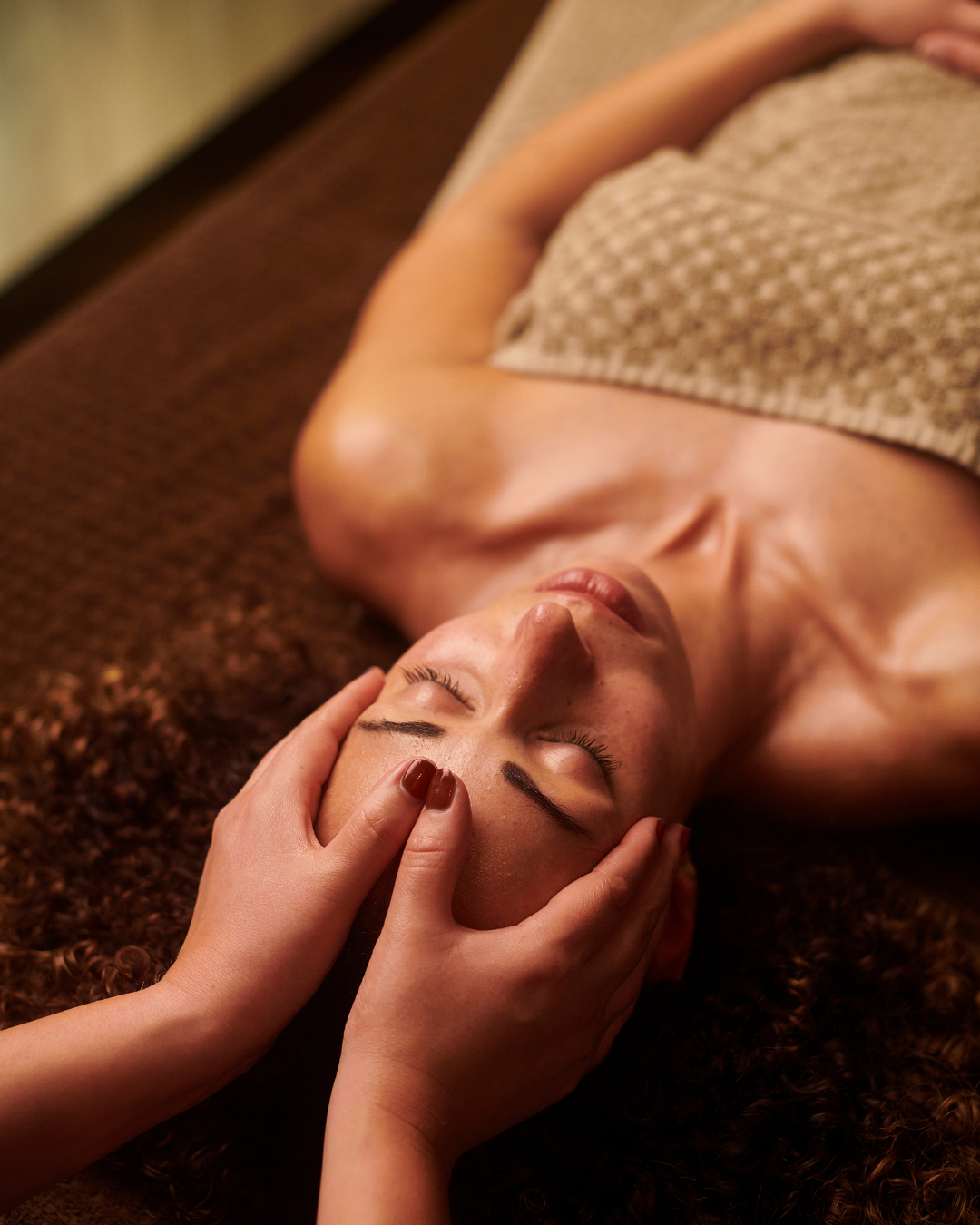 A woman lying on her back on a brown textured surface, receiving a face massage with her eyes closed, wearing a beige towel.