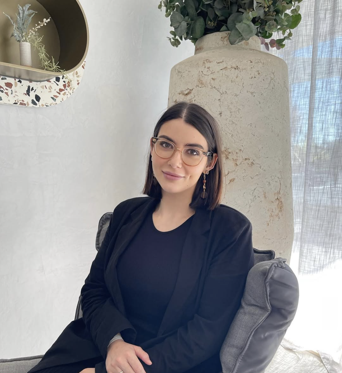 Martika Bennett, founder of Sana Skin Clinic, sits on a gray chair, wearing a black top and black blazer, in front of a large textured vase with green foliage, next to a window with sheer curtains.