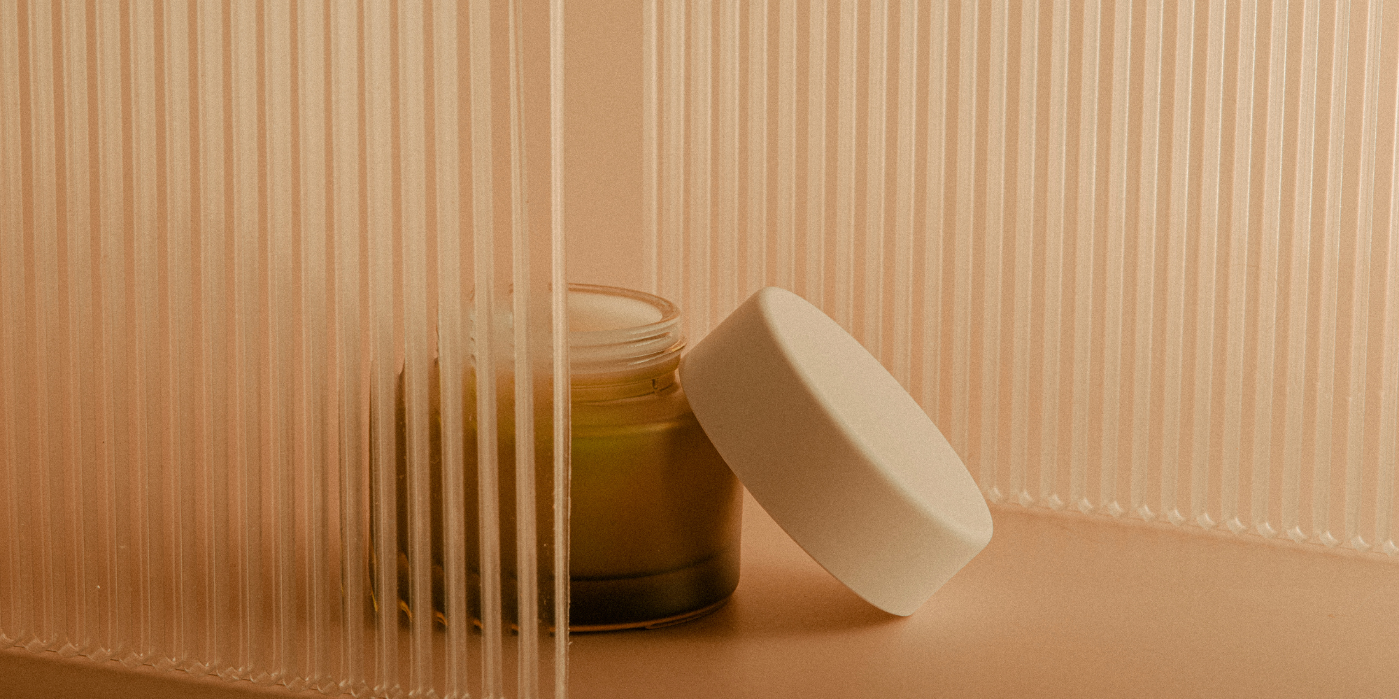 A small jar of beige cream with a smooth, rounded lid, placed on a pink surface with textured, vertical ridges in the background, partially obscured by a transparent, ribbed acrylic panel.