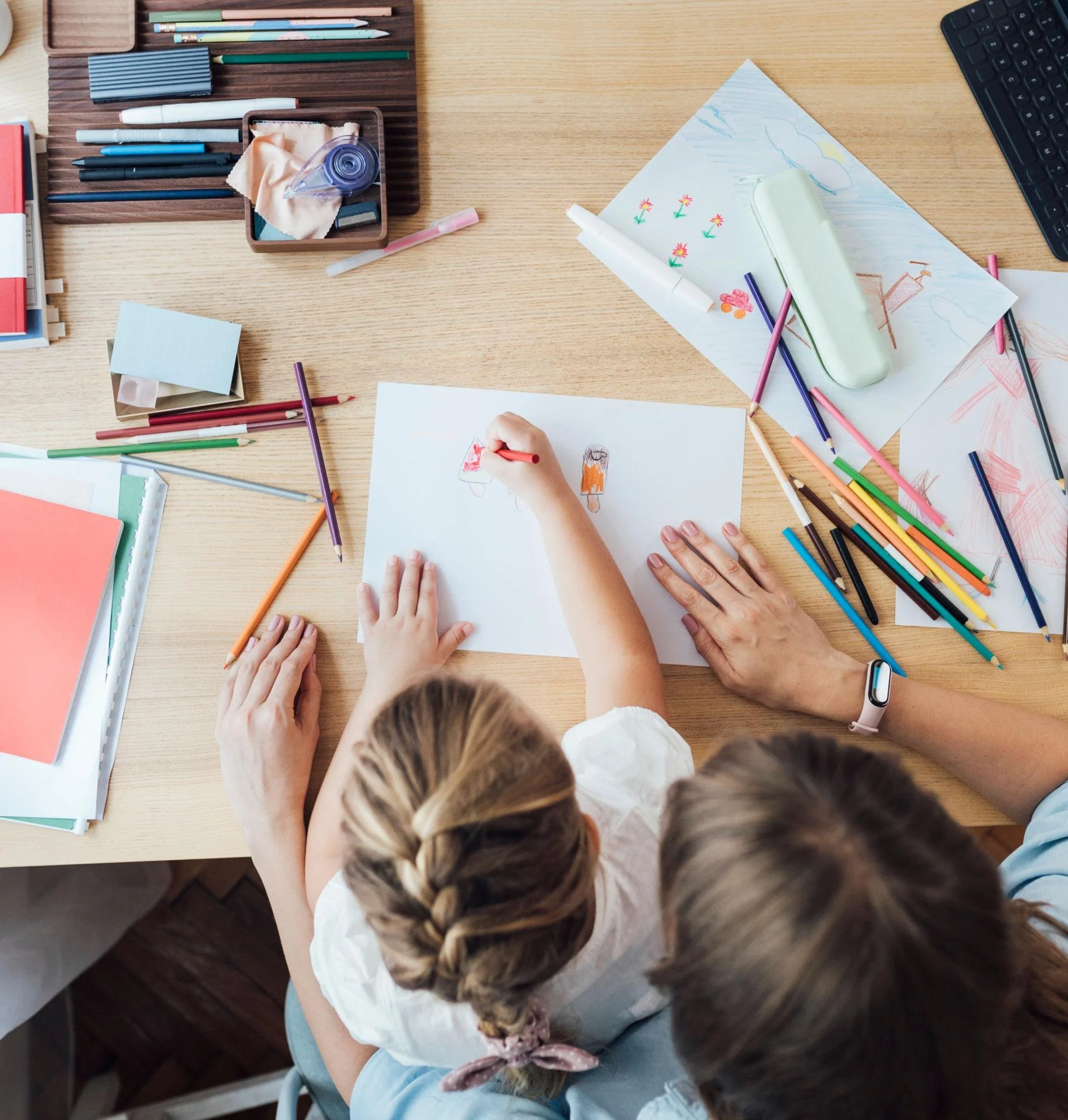 Child with braids is drawing on paper with colored pencils on a cluttered desk, which also has drawings, notebooks, and art supplies.