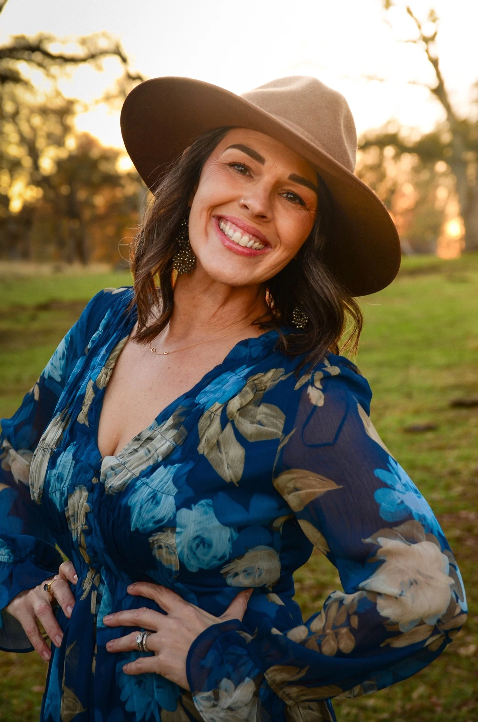 Smiling woman wearing a wide-brimmed brown hat and blue floral dress, outdoors during sunset, with trees and grass in the background.