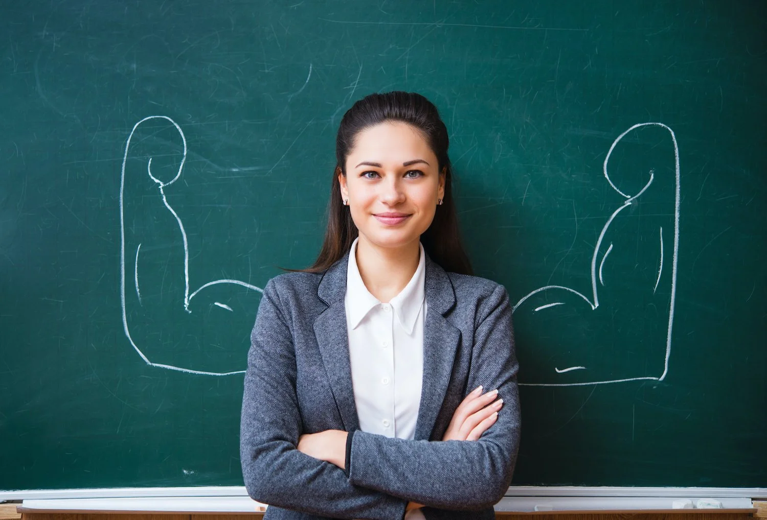 A woman standing in front of a chalkboard with white chalk outlines of muscular arms on each side of her.