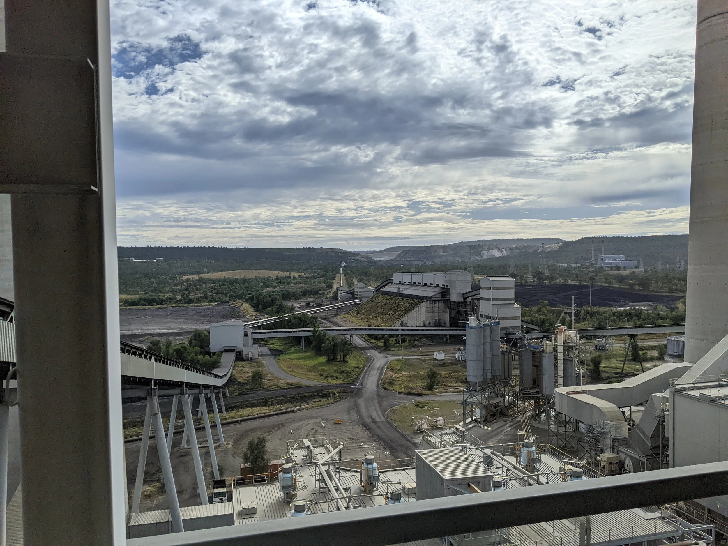 Industrial plant with large buildings, pipelines, and conveyor systems, set against a landscape with hills and cloudy sky in the background.