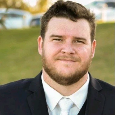 Portrait of a young man with dark hair and beard, wearing a suit jacket and tie, outdoors with blurred grass and cars in the background.