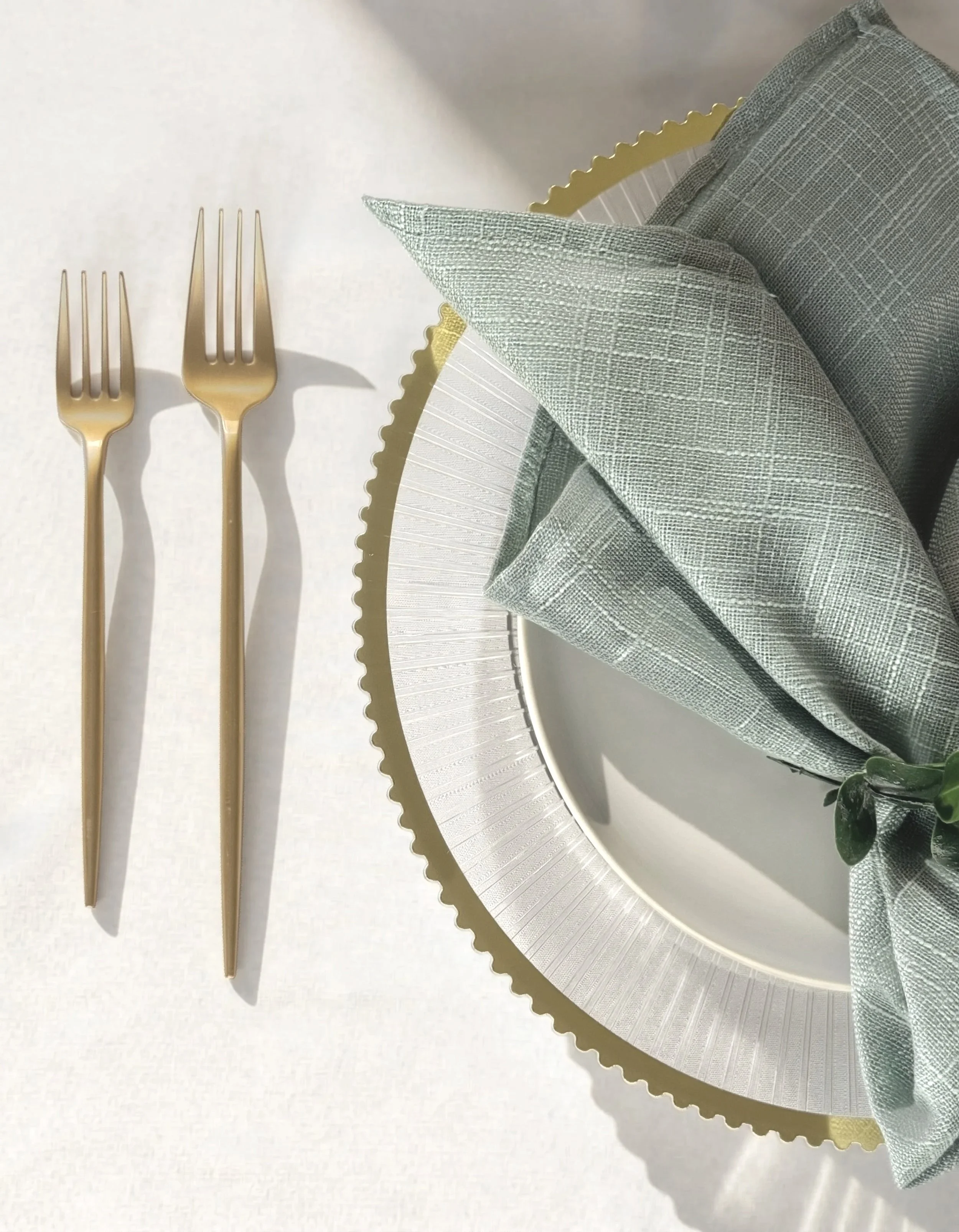 Elegant table setting with two gold forks next to a white plate with a gold scalloped edge, a folded light green napkin, and a greenery sprig on the day.