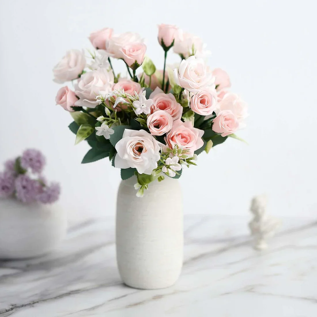 A white vase filled with pink and white roses and small white flowers on a white surface.