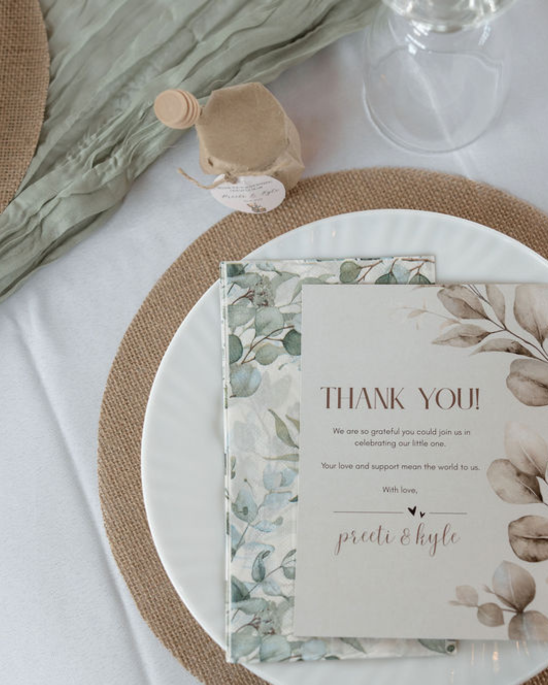 A thank you card on a white plate surrounded by a leafy napkin and a small bottle, set on a table with a burlap placemat and a glass container.