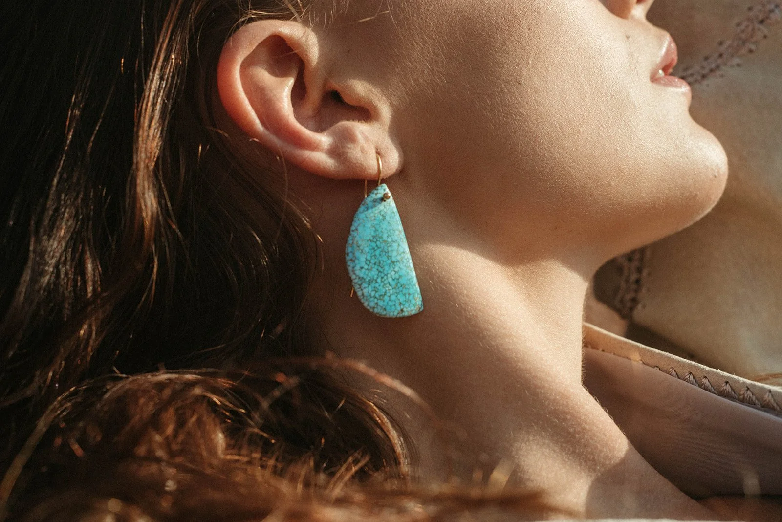 Close-up of a woman wearing a large turquoise earring, showing her ear, cheek, and part of her mouth, with sunlit skin and dark wavy hair.