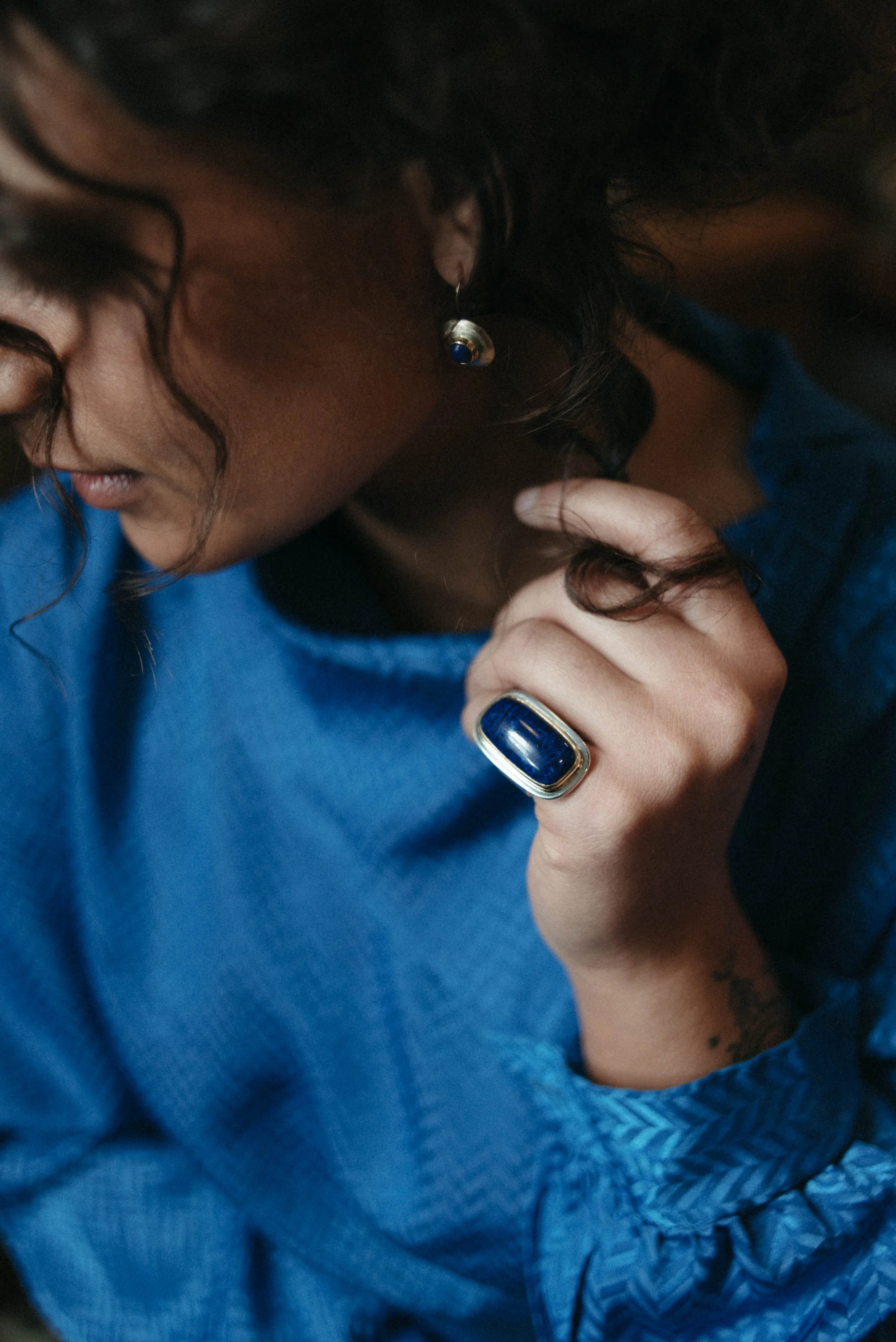 Close-up of a woman wearing a large blue ring and matching earrings, dressed in a blue top, with curly dark hair and a tattoo on her wrist.