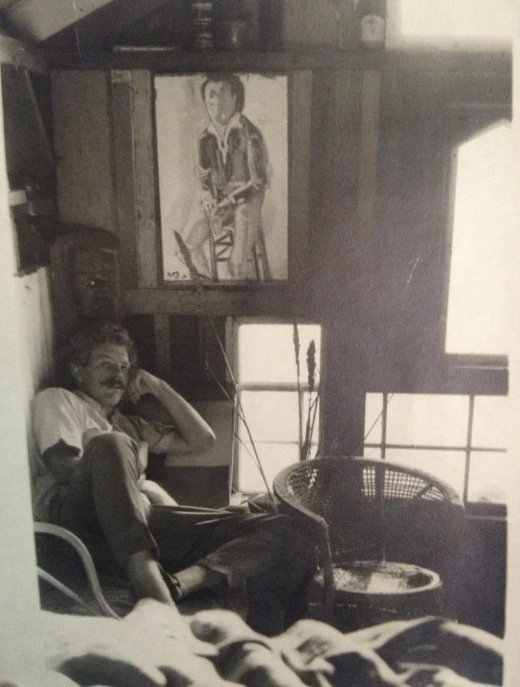 A black and white photo of a man sitting in a room with a large window and a painting of a young man hanging on the wall behind him.