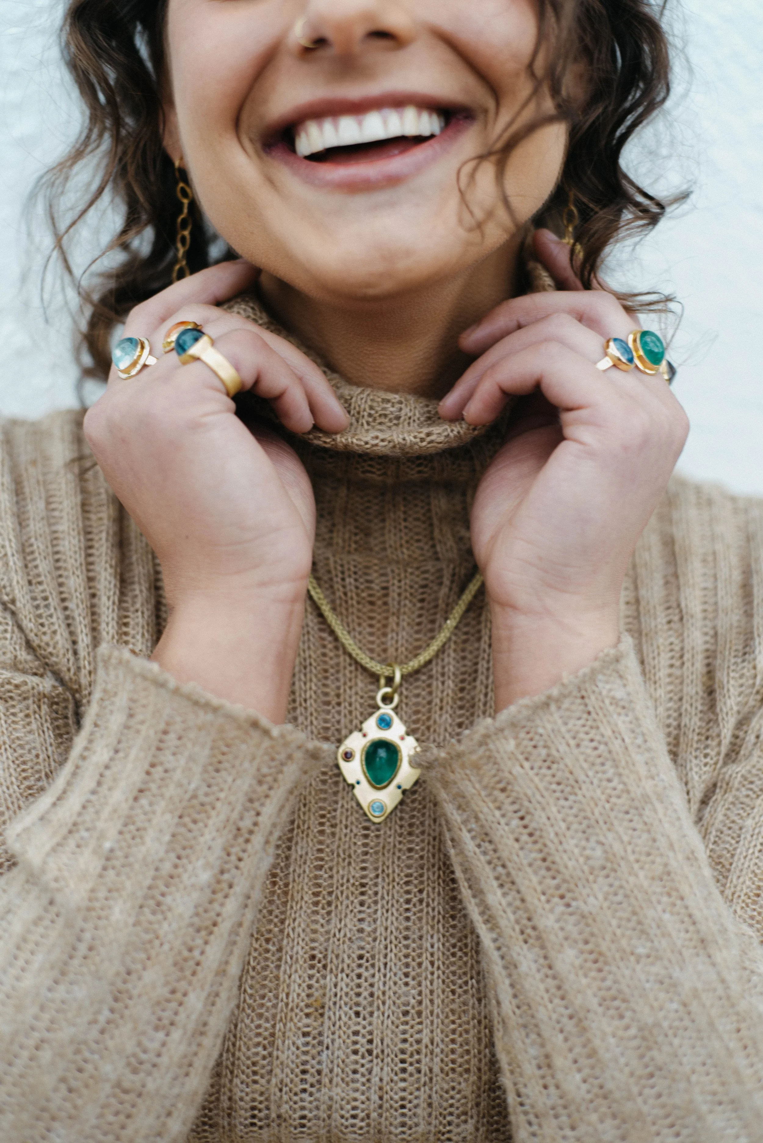 Close-up of a woman wearing a beige turtleneck sweater, with jewelry including rings, earrings, and a necklace with a large green gemstone pendant. She is smiling and holding her hands near her face.