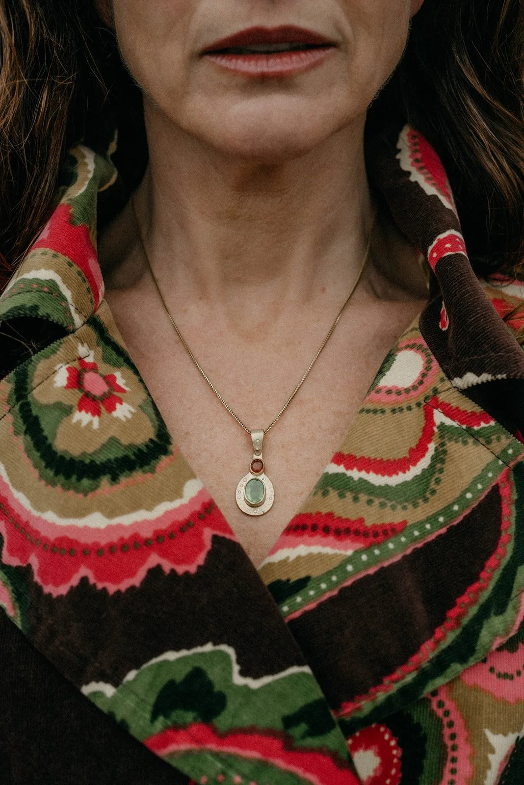 Close-up of a woman's neck and chest area, showing a gold necklace with a pendant. She wears a colorful patterned scarf with floral and geometric designs in red, green, black, and white.