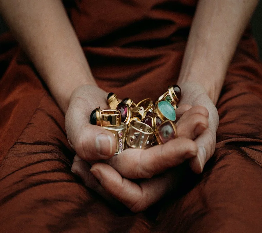 Person holding a collection of gold rings with various gemstones at their lap, wearing a rust-colored garment.