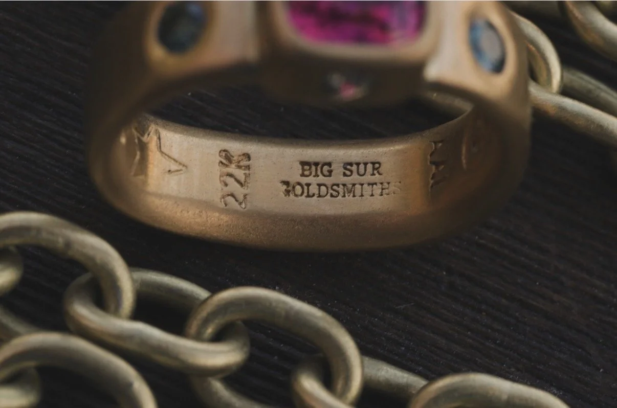 Ali Lee's ring with engravings inside reading 'BIG SUR GOLDsMITH' and the number '22K', surrounded by metal chains on a black surface.