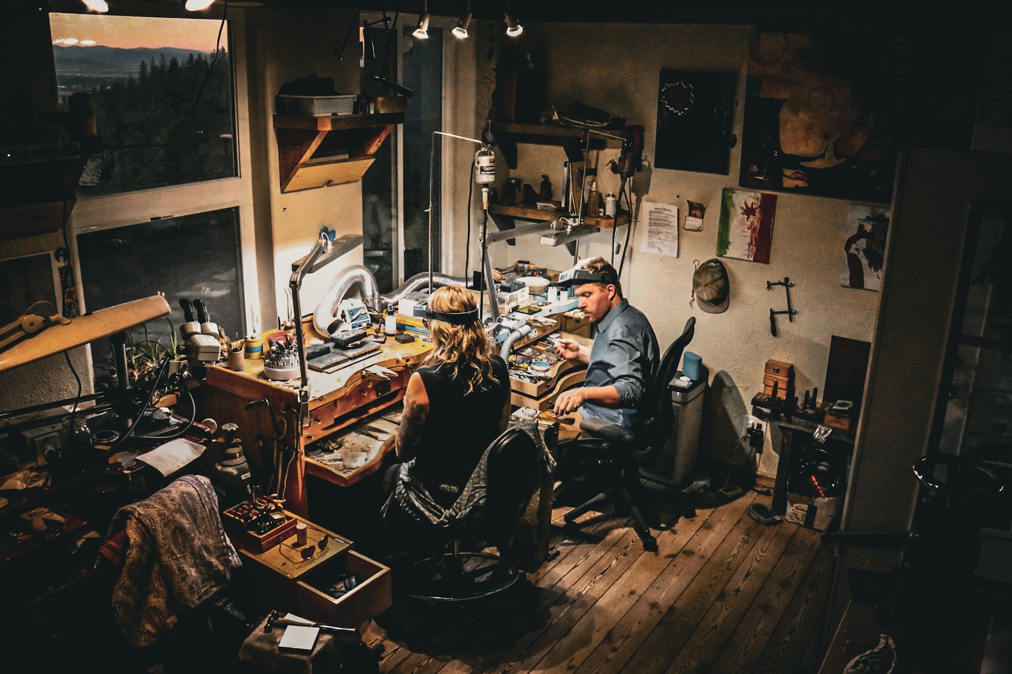 A photograph of Ali and Carrie working in their jewelry studio, taken by their daughter Beatriz, with a view of mountains visible through the window in the background.