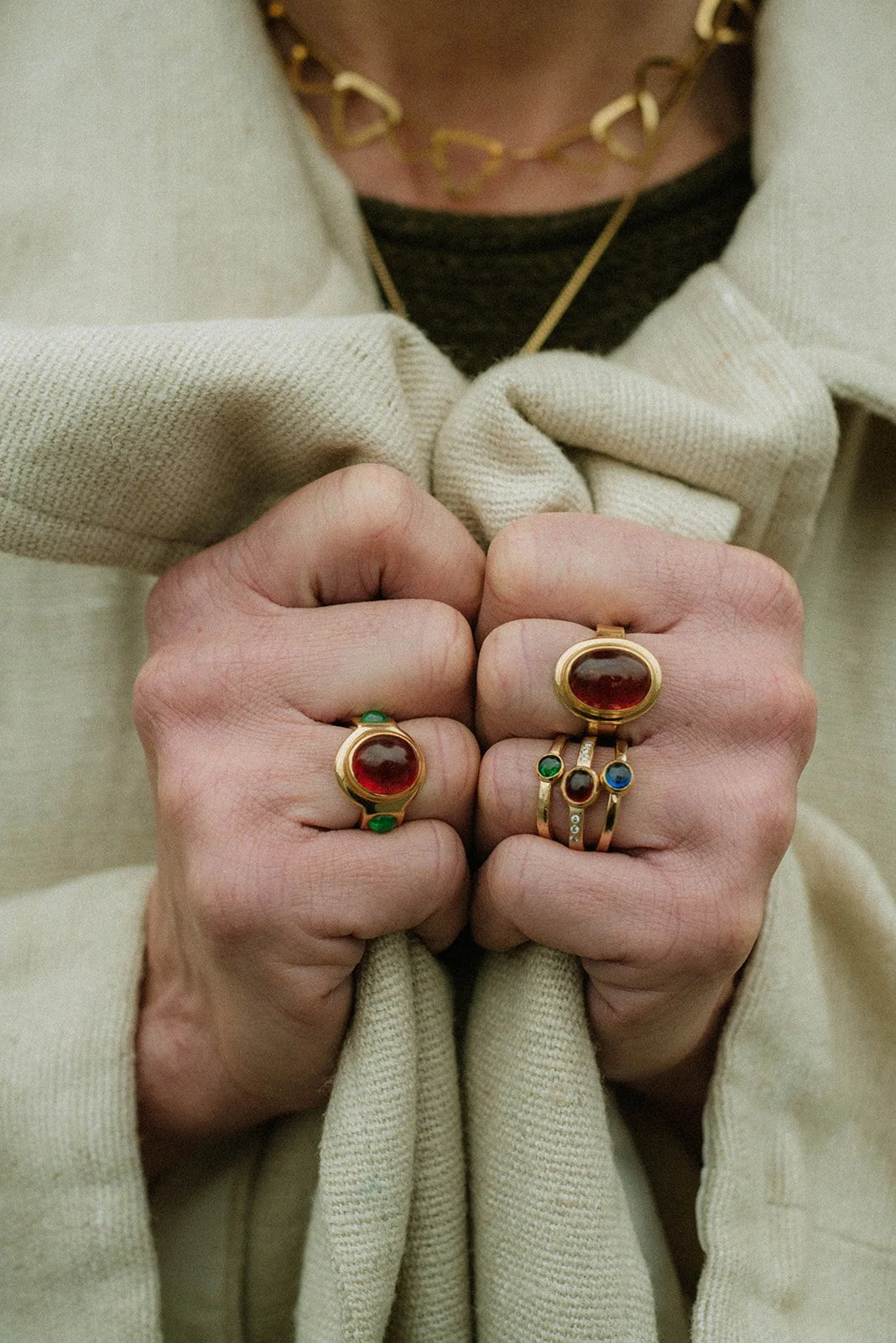 Close-up of a person wearing multiple rings with large red stones, gold bands, and small green, blue, and diamond accents, gripping a beige fabric with a gold chain necklace visible around the neck.